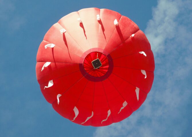 View looking up at a hot air balloon against a blue sky. The hot air balloon is bright red.