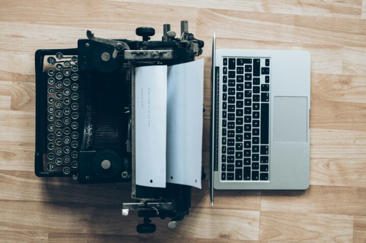 A typewriter sitting on a desk, back-to-back with a laptop